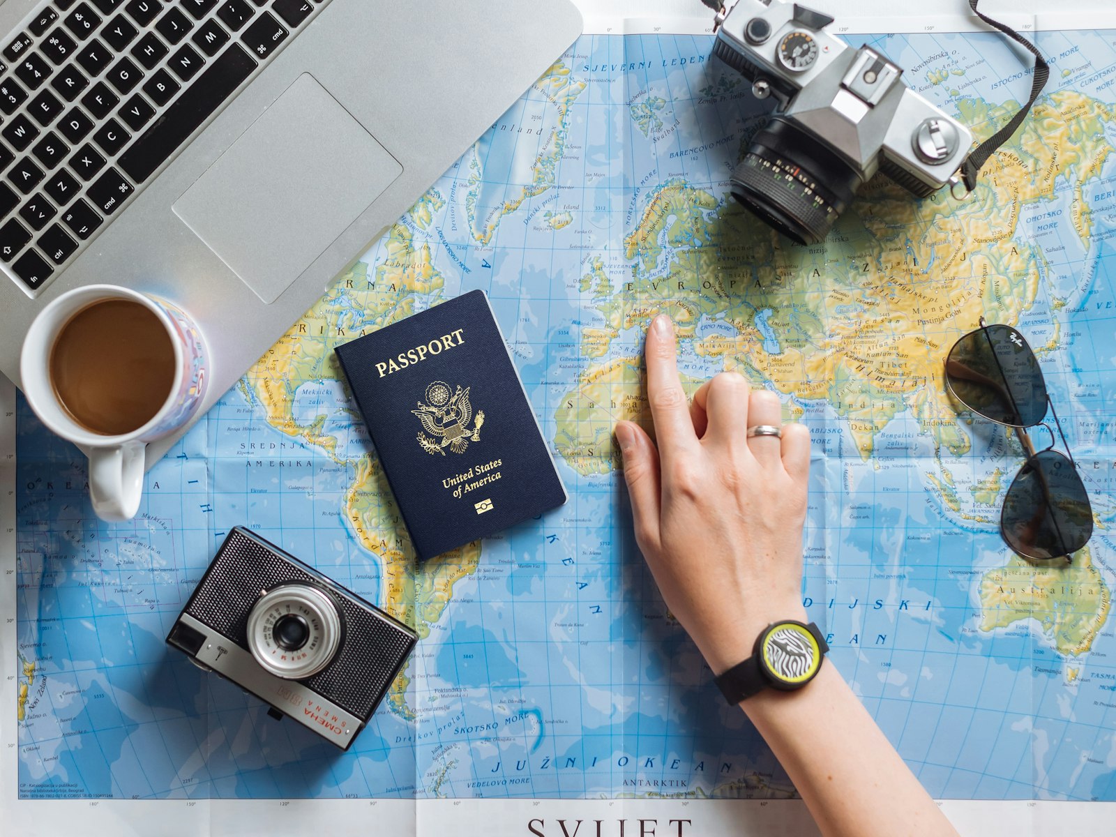 Mano señalando un destino en un mapa del mundo, con pasaporte, cámaras y un portátil